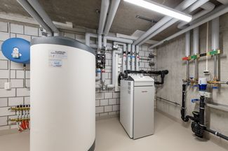 Technikraum mit einem grossen Warmwasserspeicher, einer modernen Heizungsanlage und sauber verlegten Rohrleitungen sowie verschiedenen Armaturen und Regelungskomponenten an der Wand.