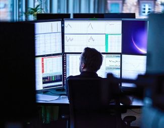 Ein Mann sitzt im dunklen Tradingfloor der BKW vor sechs grossen Bildschirmen. Darauf sind verschiedene Grafiken und Tabellen abgebildet.