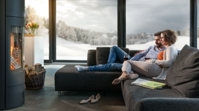 Personen sitzen entspannt auf einem modernen, dunklen Sofa vor einem knisternden Kaminfeuer, mit Blick auf eine verschneite Landschaft durch grosse Panoramafenster. Ein Korb mit Holz steht neben dem Kamin, Zeitschriften und Schuhe liegen im Wohnbereich.