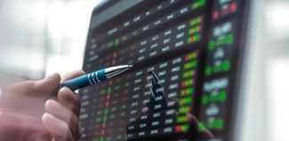 Auf einem Computerbildschirm sind viele Zahlen abgebildet, einige sind rot oder grün hinterlegt. Eine Hand hält einen Kugelschreiber, der auf den Bilschirm zeigt. 